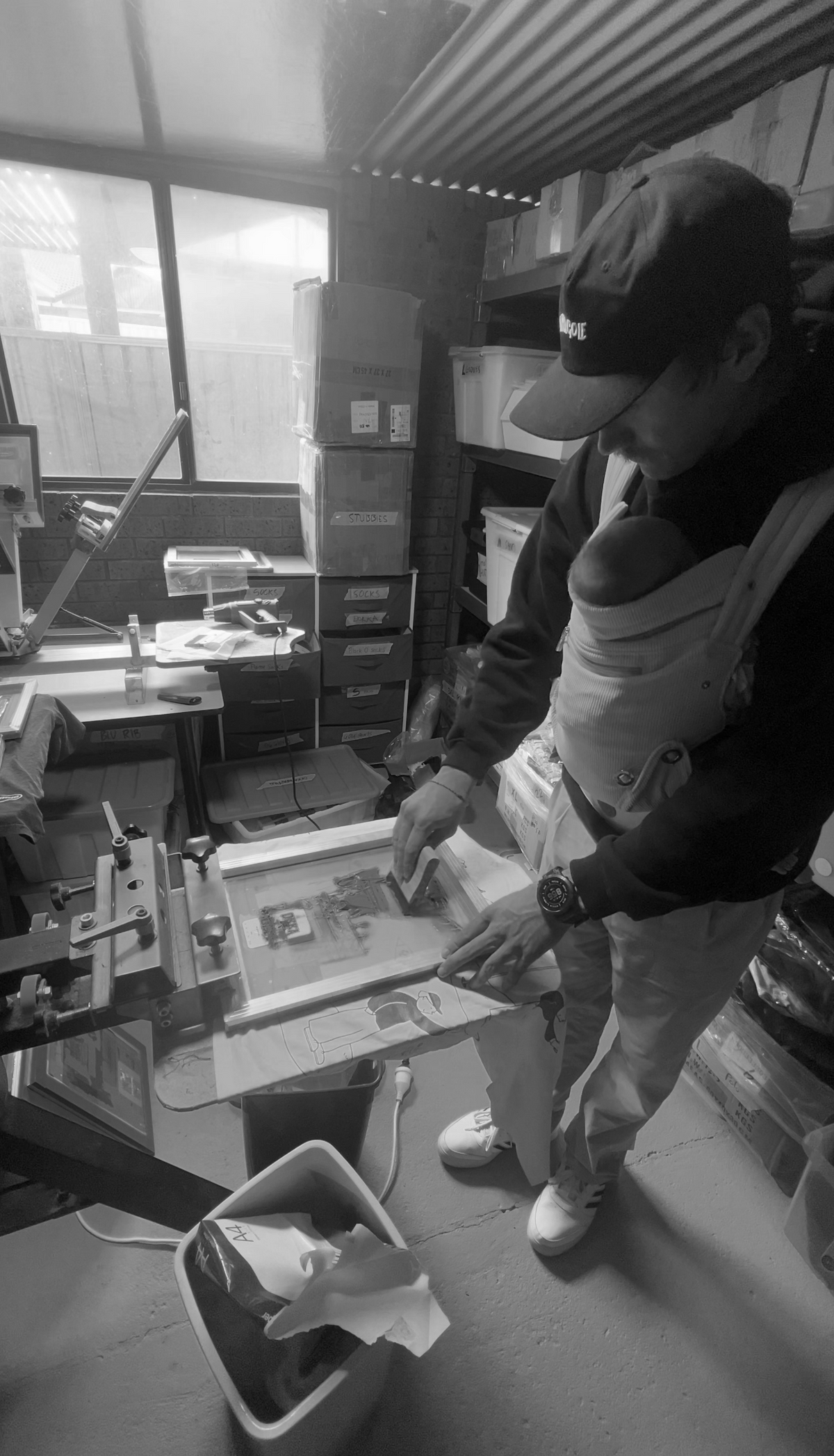 A person in a baby carrier stands in a workshop or studio, holding a printed sheet near a screen printing machine. The scene is shown in black and white, with shelves, boxes, and equipment visible in the background.