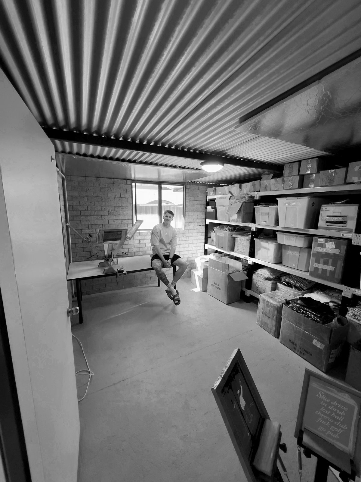 A black and white photo of a man sitting on a chair in a storage room with shelves of boxes, a corrugated metal ceiling, and a brick wall.